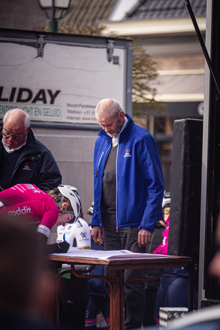 An older man in a blue jacket stands at a table with people on a street near a sign that reads "Milday".