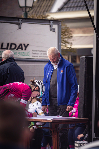 A man in a blue jacket stands near cyclists in front of a van with the word Wielrennen.