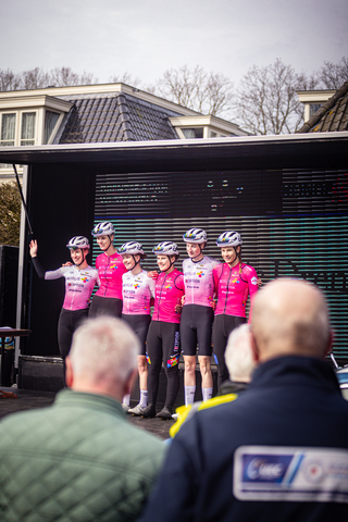 A group of cyclists wearing pink and white uniforms pose for a photo with the audience in the background.