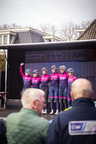 A group of people wearing cycling outfits stand together on stage, with a person raising their hand.