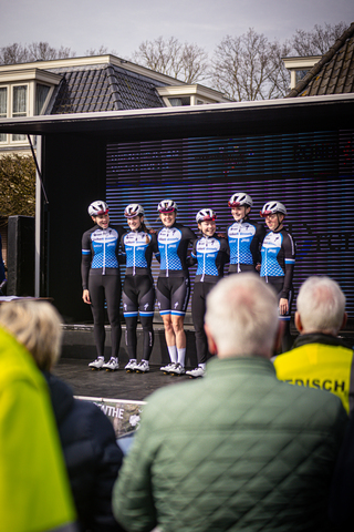 A group of cyclists stand on a stage in front of a large screen, with spectators looking on.