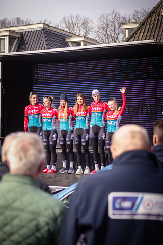 A group of female cyclists are standing in a row on a stage, all wearing matching blue and red uniforms.