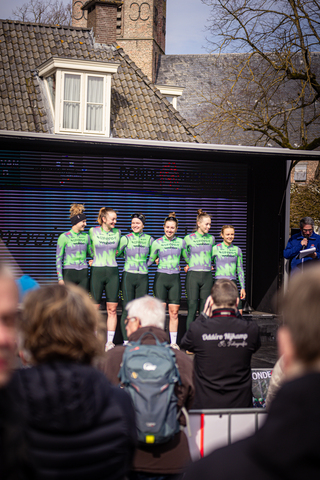 Several young women wearing Wielrennen jerseys stand together in front of a black screen.