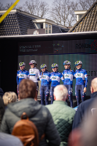 A group of cyclists stand for a photo in front of a large screen with the words "Drentse 8 van Westerveld" on it.