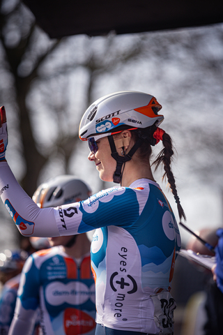 A cyclist wearing a blue and white jersey with the word "drentse" on it waves to someone.