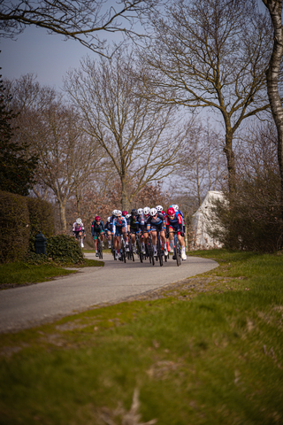 A group of cyclists race on a path near trees, a white tent, and a blue water tank.