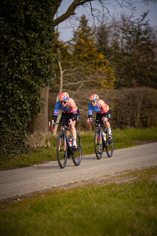 Two cyclists with helmets of different colors are wearing matching team colors and are riding their bikes down a narrow road.