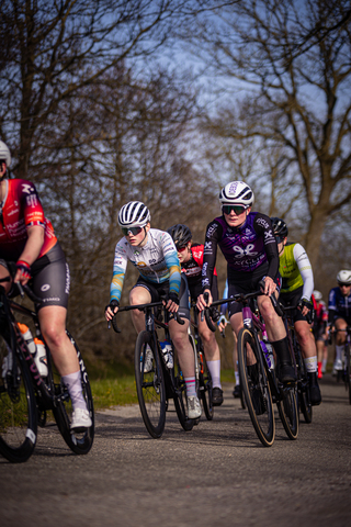 Eight cyclists, including one woman in purple and another man in black, race on a road lined with trees.