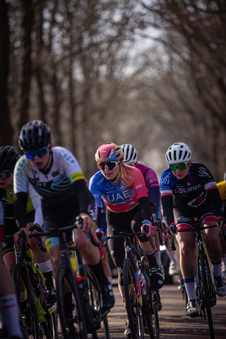 A group of cyclists are wearing helmets and riding their bikes on a road, with one cyclist in the foreground.