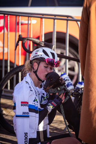 A woman wearing a white Adidas jersey is petting a dog with her face near the dog's head.