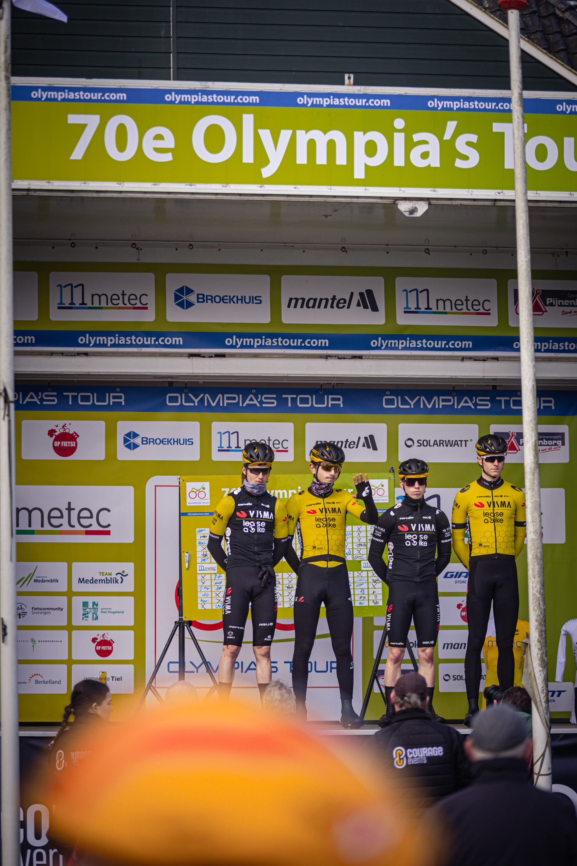 A group of cyclists are standing in front of a blue and green sign that reads "70e Olympia's Tour".