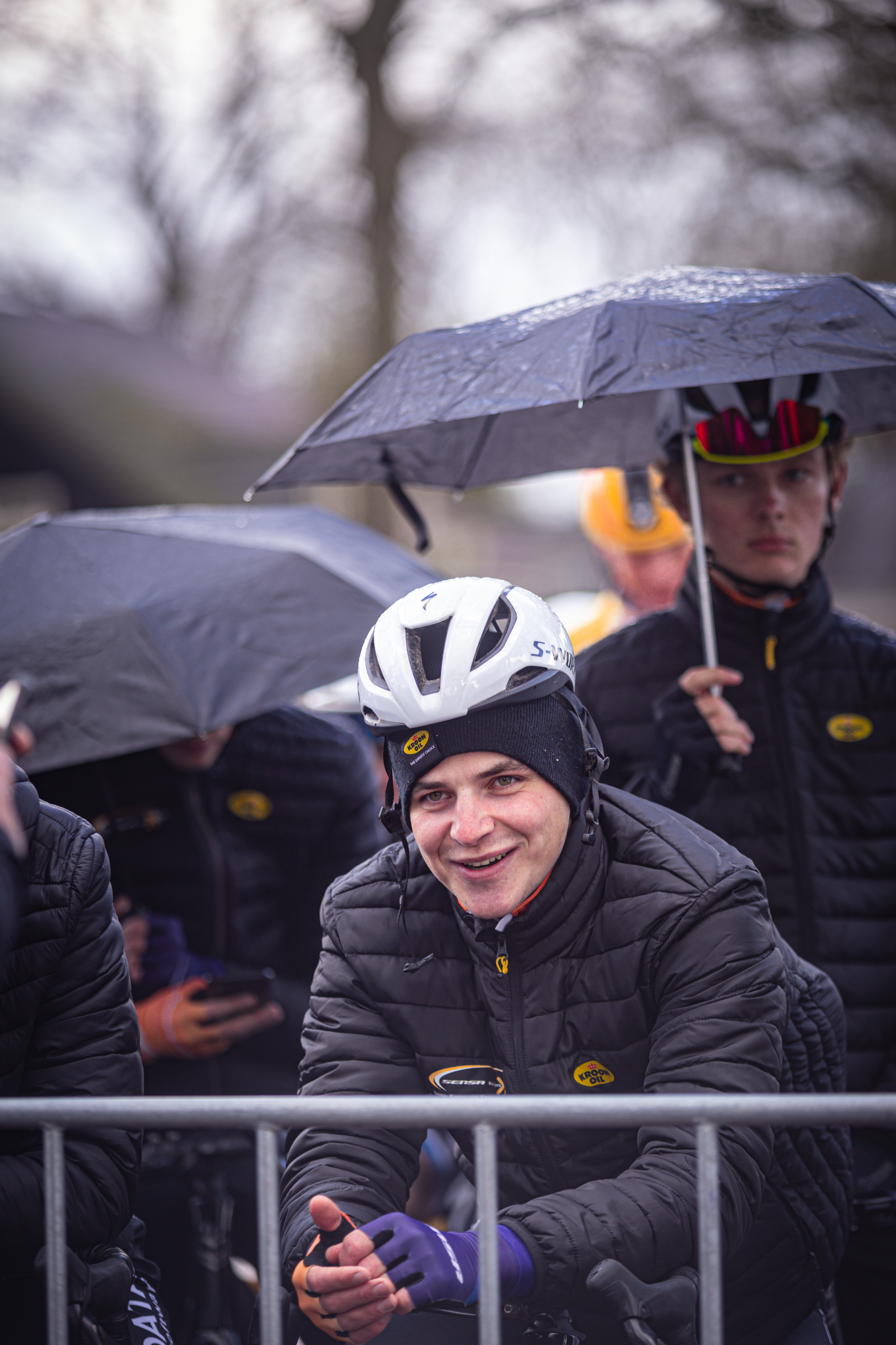 A man wearing a helmet smiles as he holds an umbrella in front of a crowd at the Olympics.