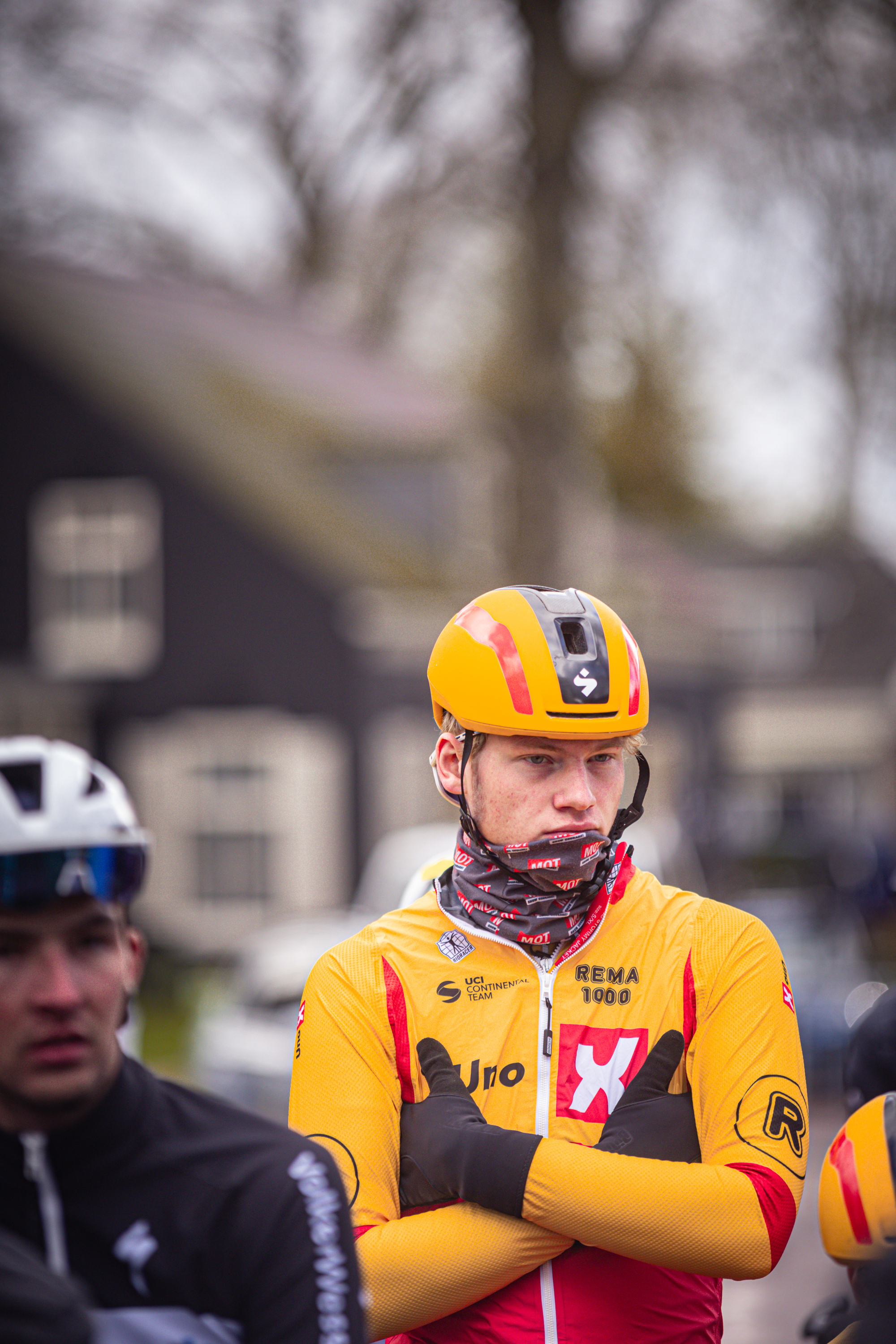 Two cyclists, one wearing a red and yellow shirt with the word "JOMA" written on it, stand next to each other.