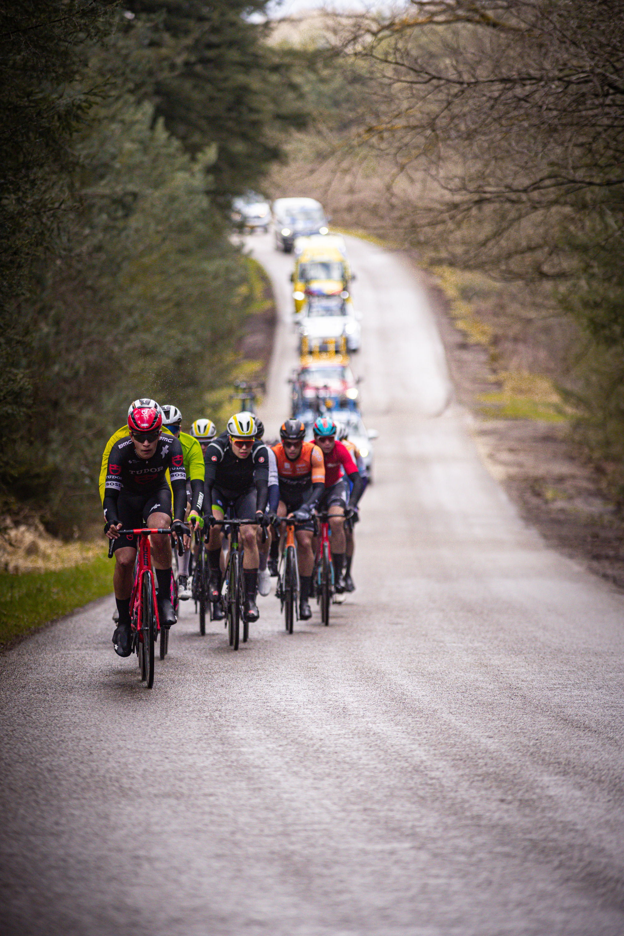 A group of cyclists race on a country road, wearing helmets and riding red and yellow bikes.