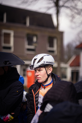 A young man in a white and black helmet is wearing a jacket with the letters "S" and "Wielrennen". He is looking up.