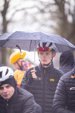 A young cyclist with a helmet and an umbrella stands next to another cyclist, both wearing black jackets.