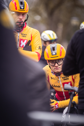 Group of cyclists at the Tour de France, wearing yellow and orange uniforms with their helmets visors down as they ride.