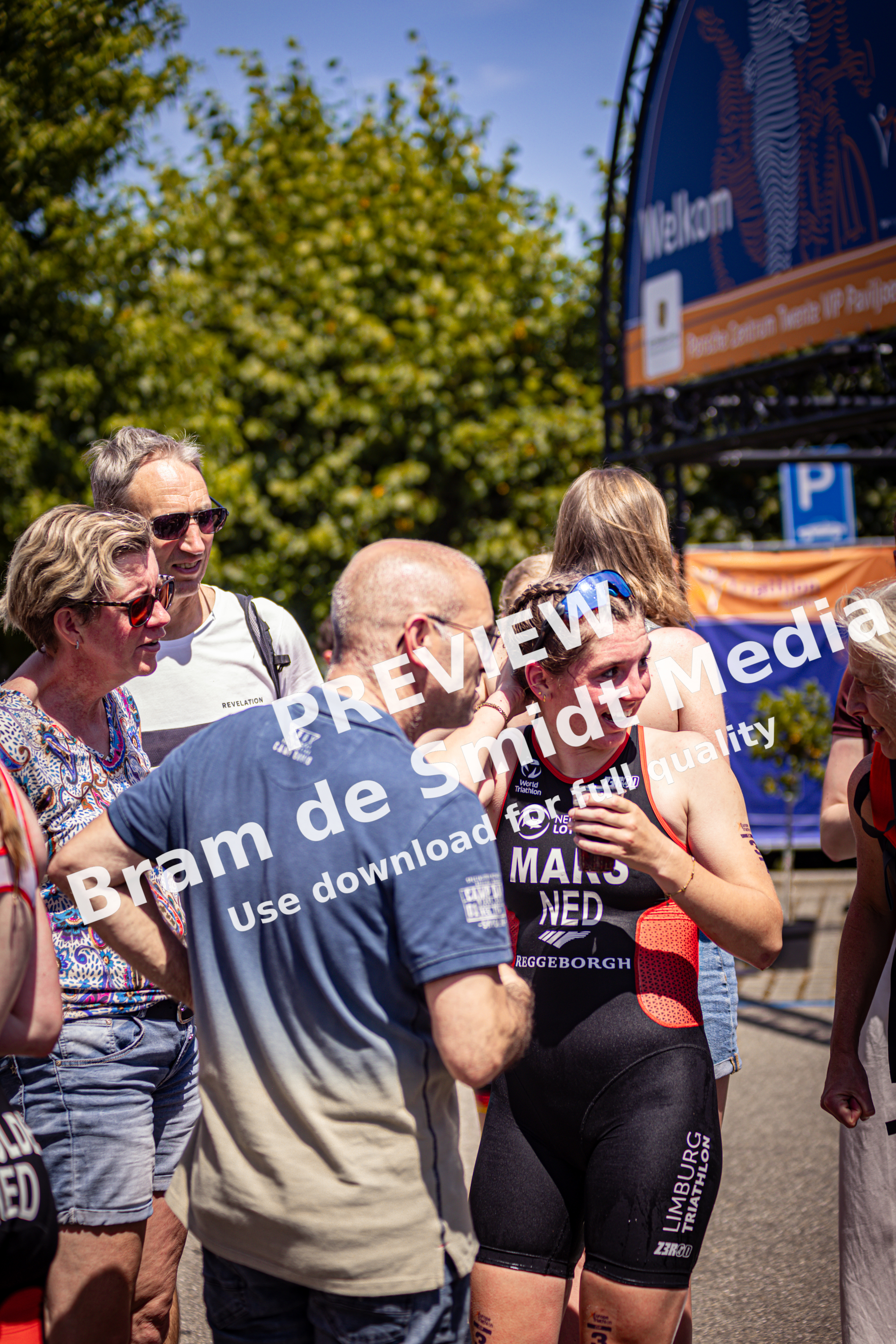 A woman is wearing a blue headband and an Elite Triathlon Unit shirt, standing near a group of people.