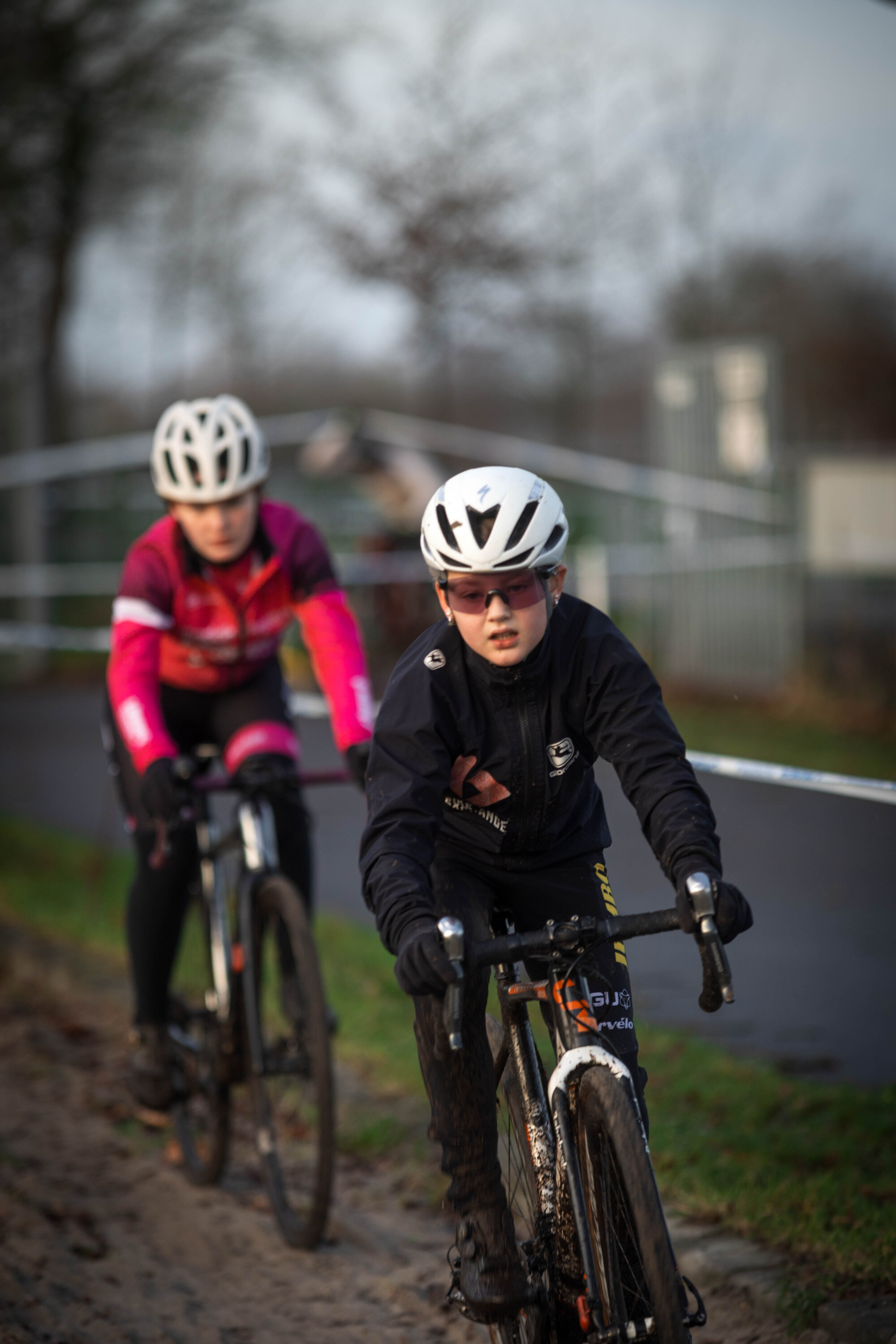 Two young cyclists are on a path, one wearing a white and black helmet and the other in pink.