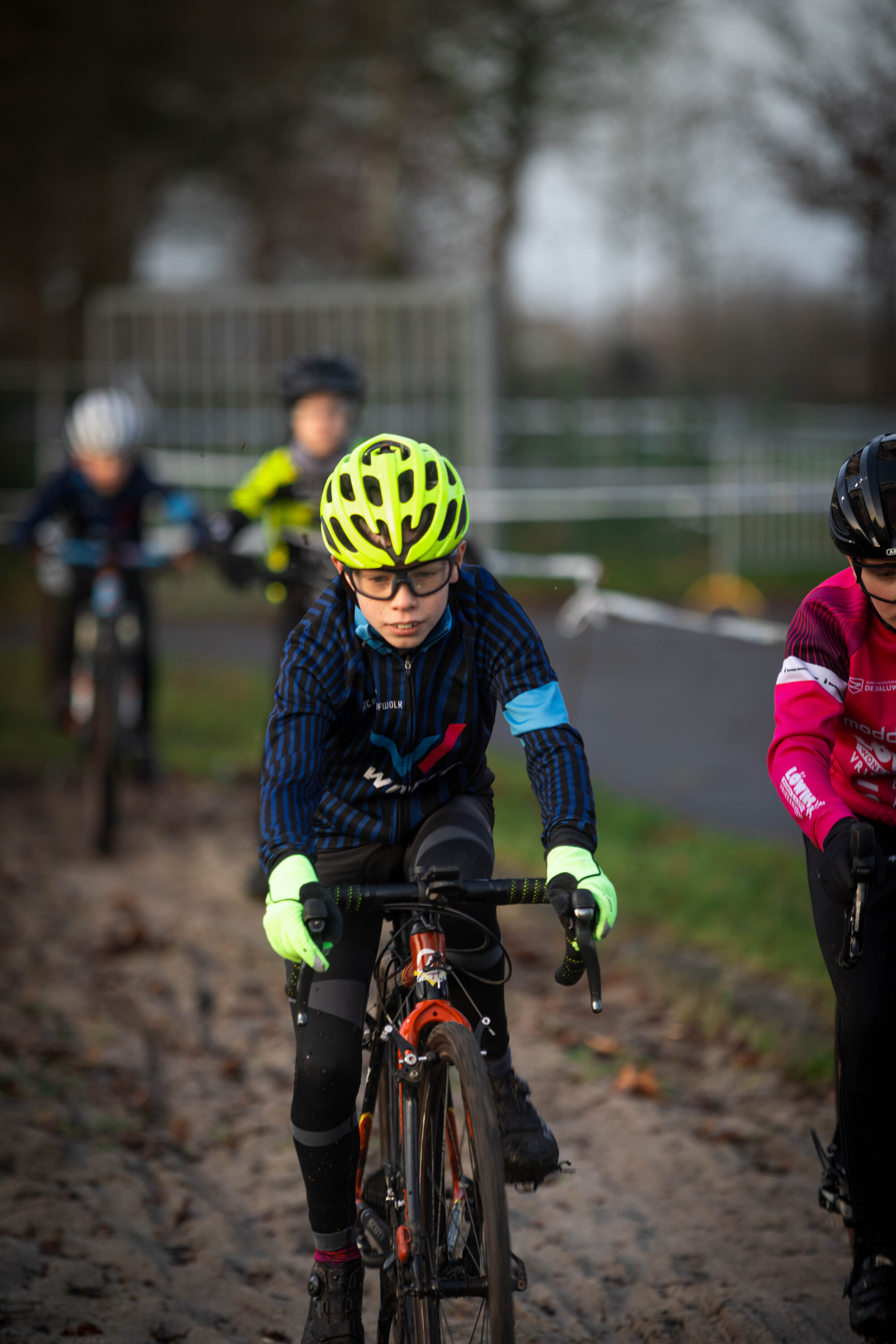 Jeug cyclocrossers are racing on a dirt track, wearing safety helmets and gloves.