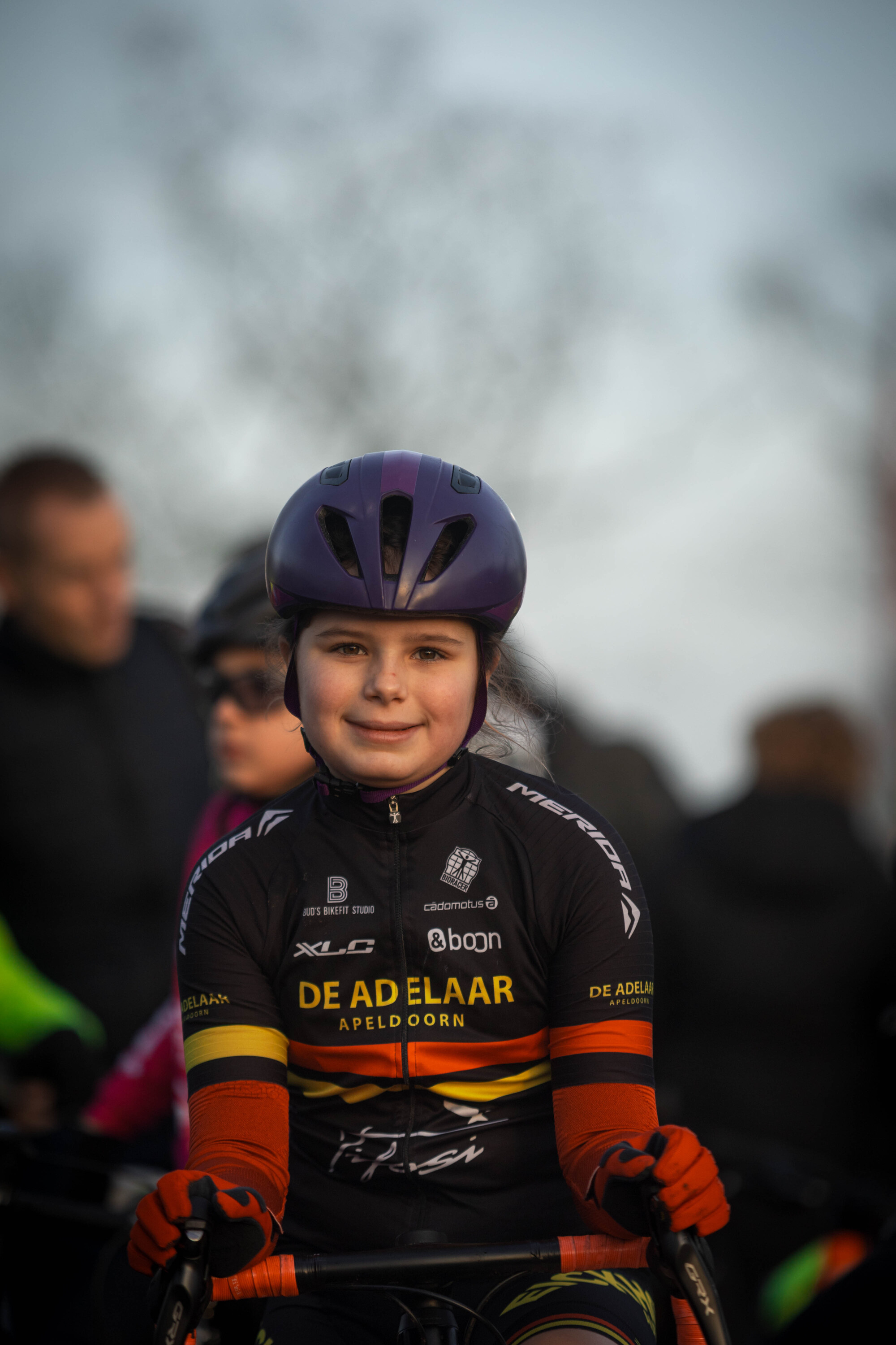 A young girl is wearing a purple helmet and black jersey that says "De Adelaar" and "Lysen".