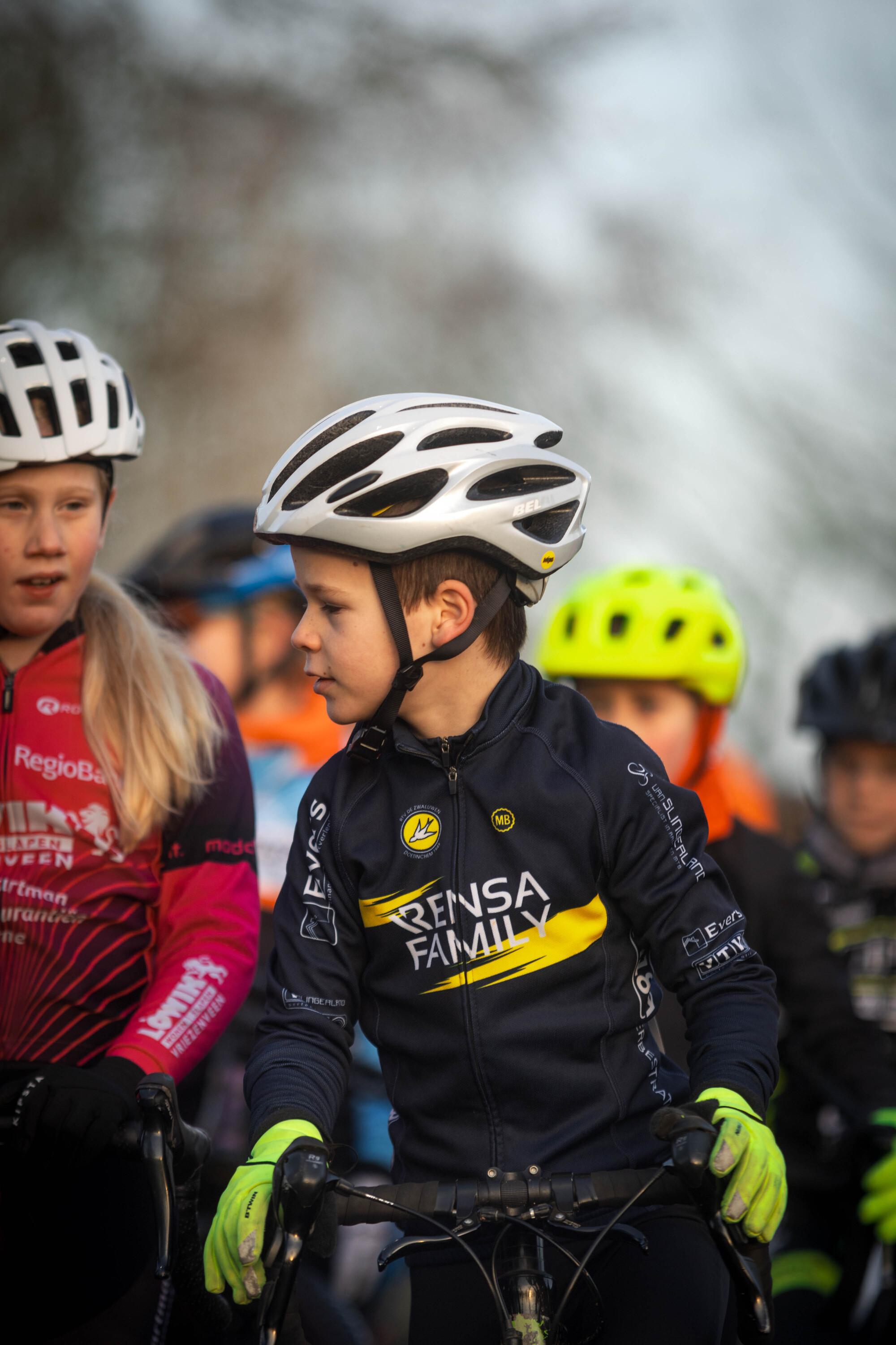 A child wearing a black jersey with the words "Genss Family" on it, stands next to other children in cycling gear.