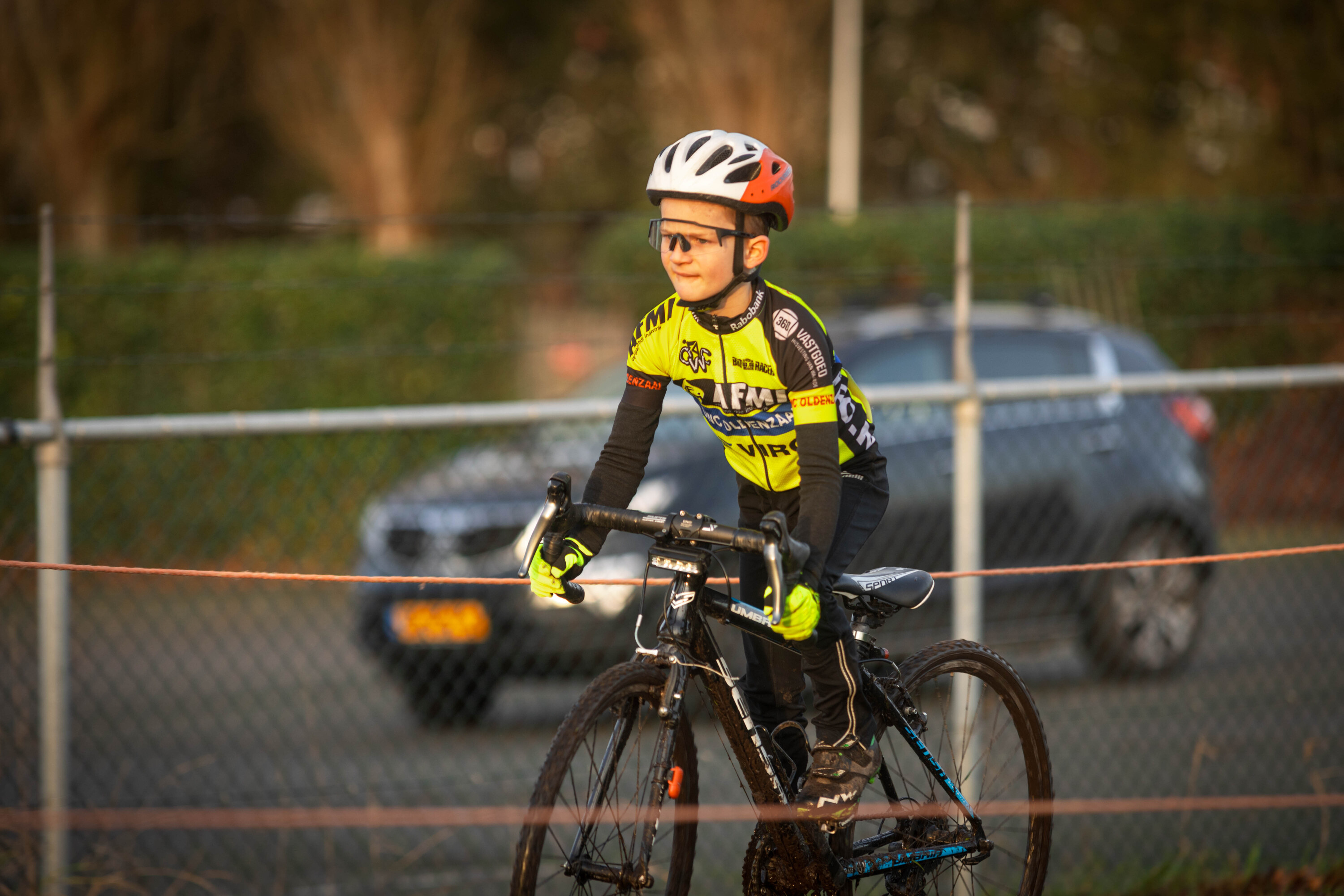 A child wearing a yellow and black shirt is riding a bike near a car.