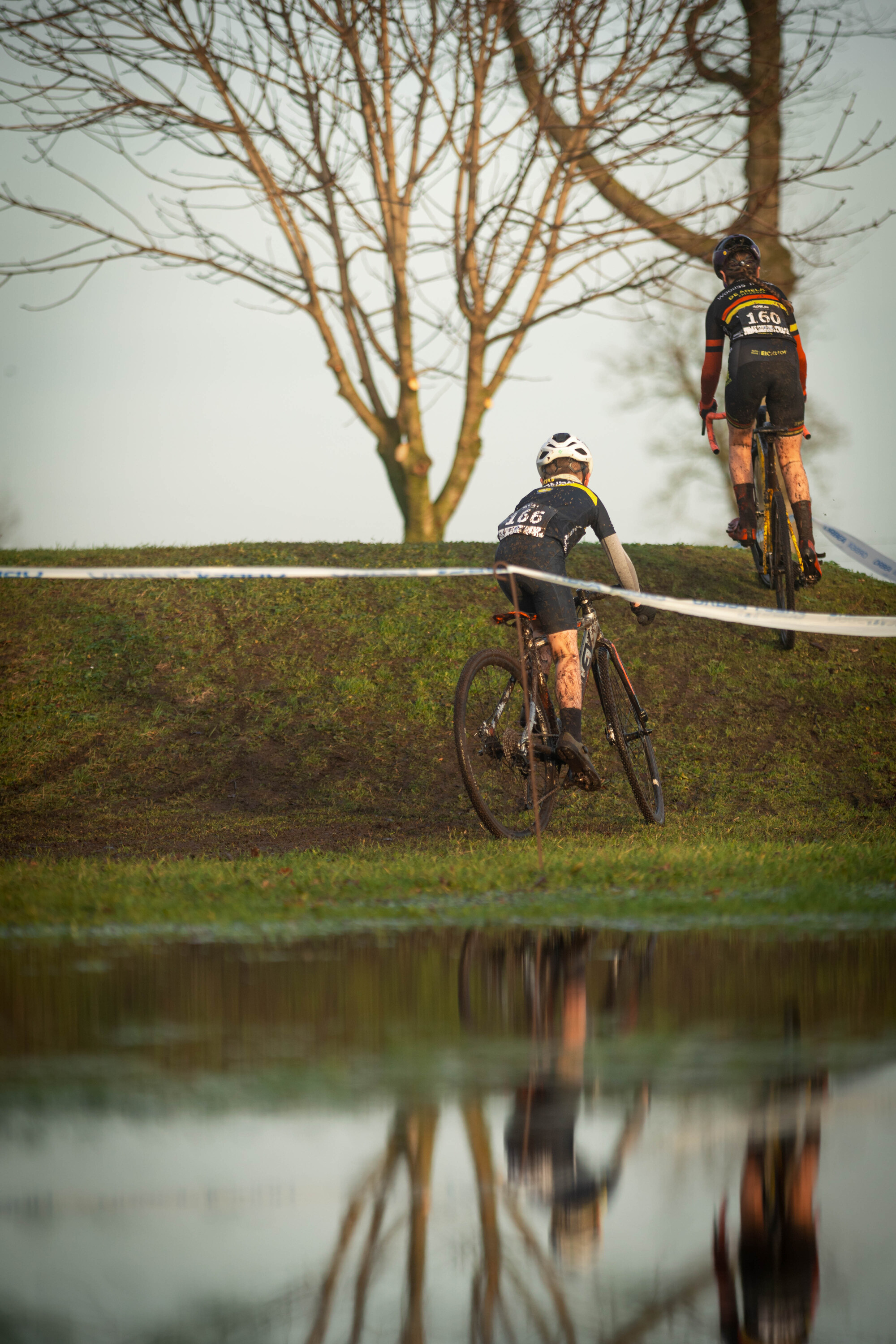 Two young cyclists wearing black and white uniforms are riding their bikes on a grassy hill, racing in a cyclocross race.