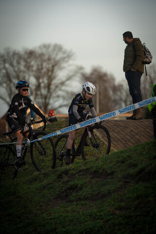 Two cyclists on a dirt hill with a third man standing nearby.