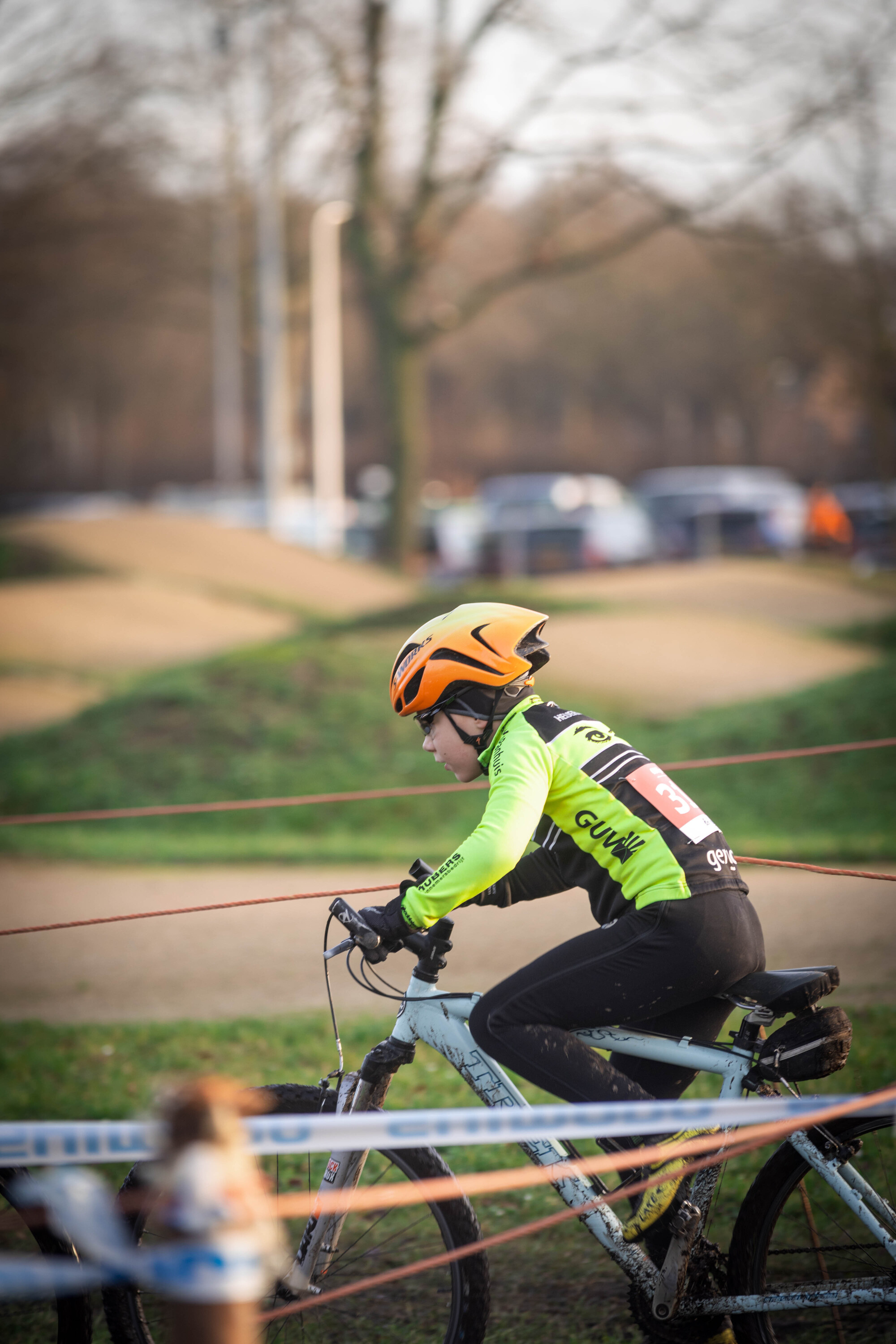 A young person wearing a yellow helmet and a green shirt with the word Jeug on it is riding a blue bicycle.