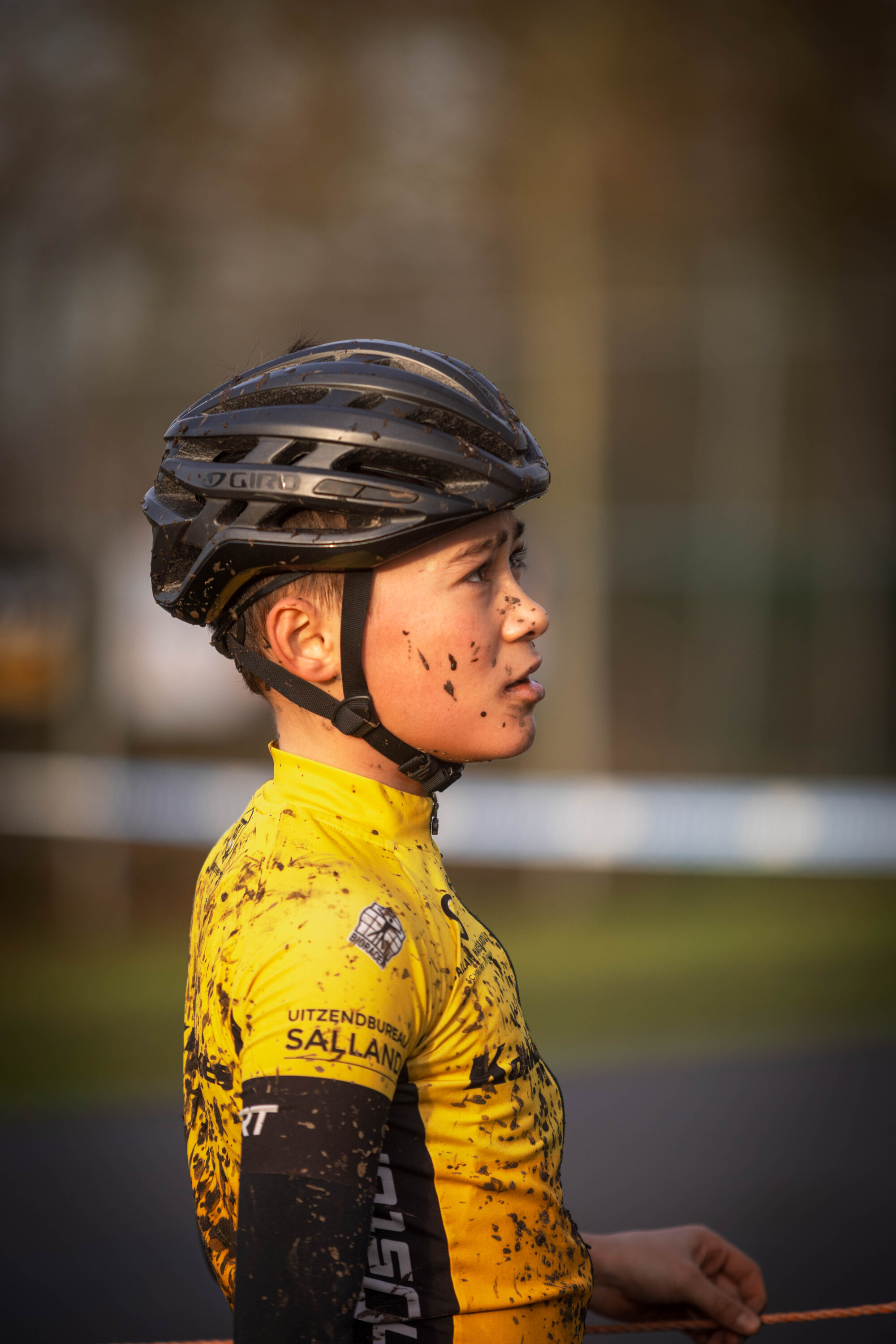 A young woman in a yellow jersey is wearing a black bike helmet and standing by the road.