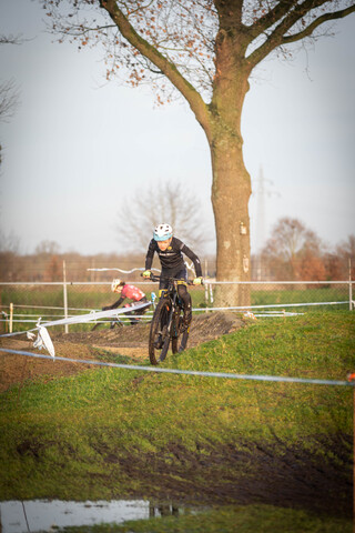 A man is riding a bike on a dirt track with a white dog standing nearby.