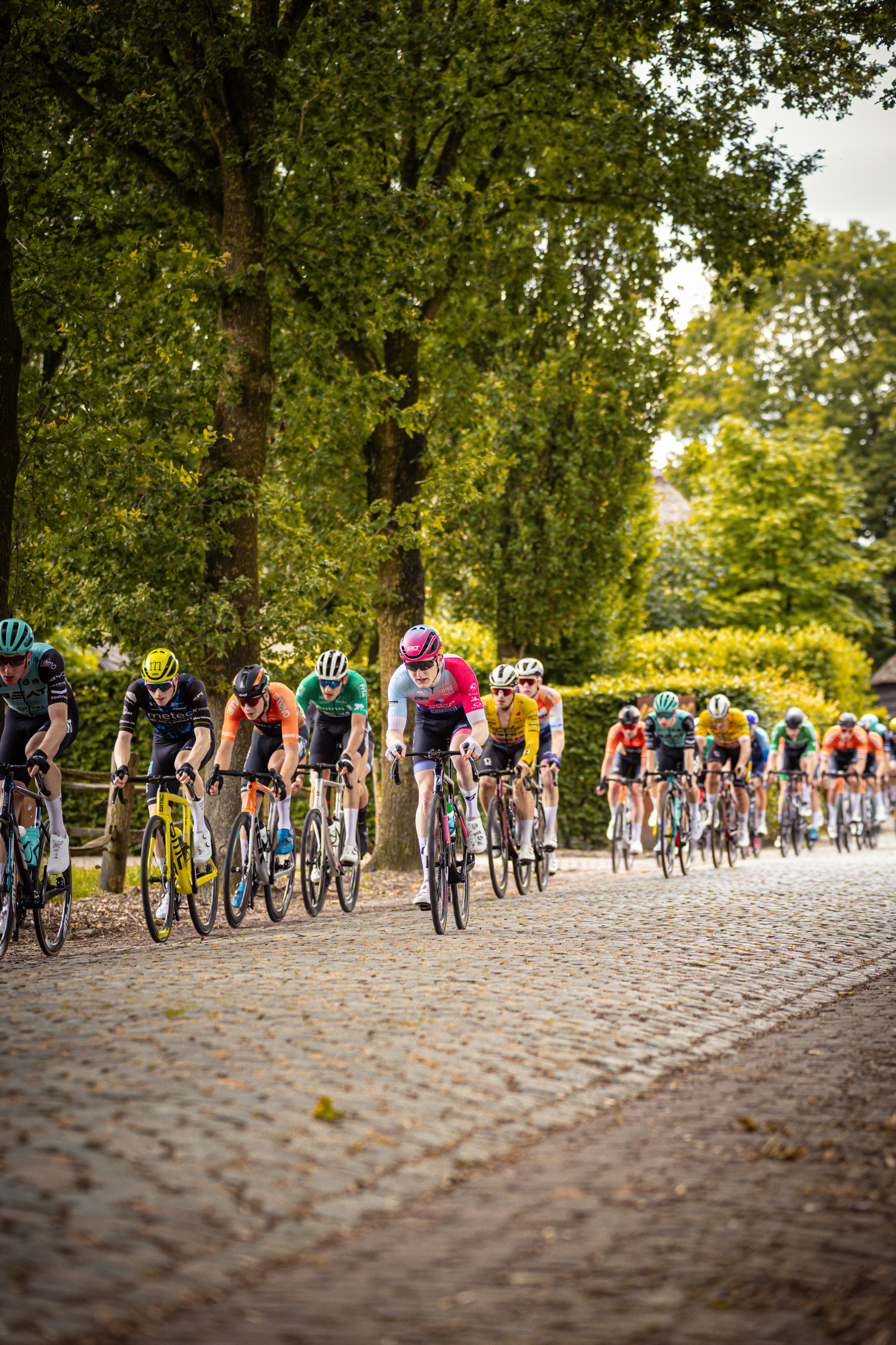 A group of cyclists on a cobblestone road with Midden Brabant Poort Omloop in the background.