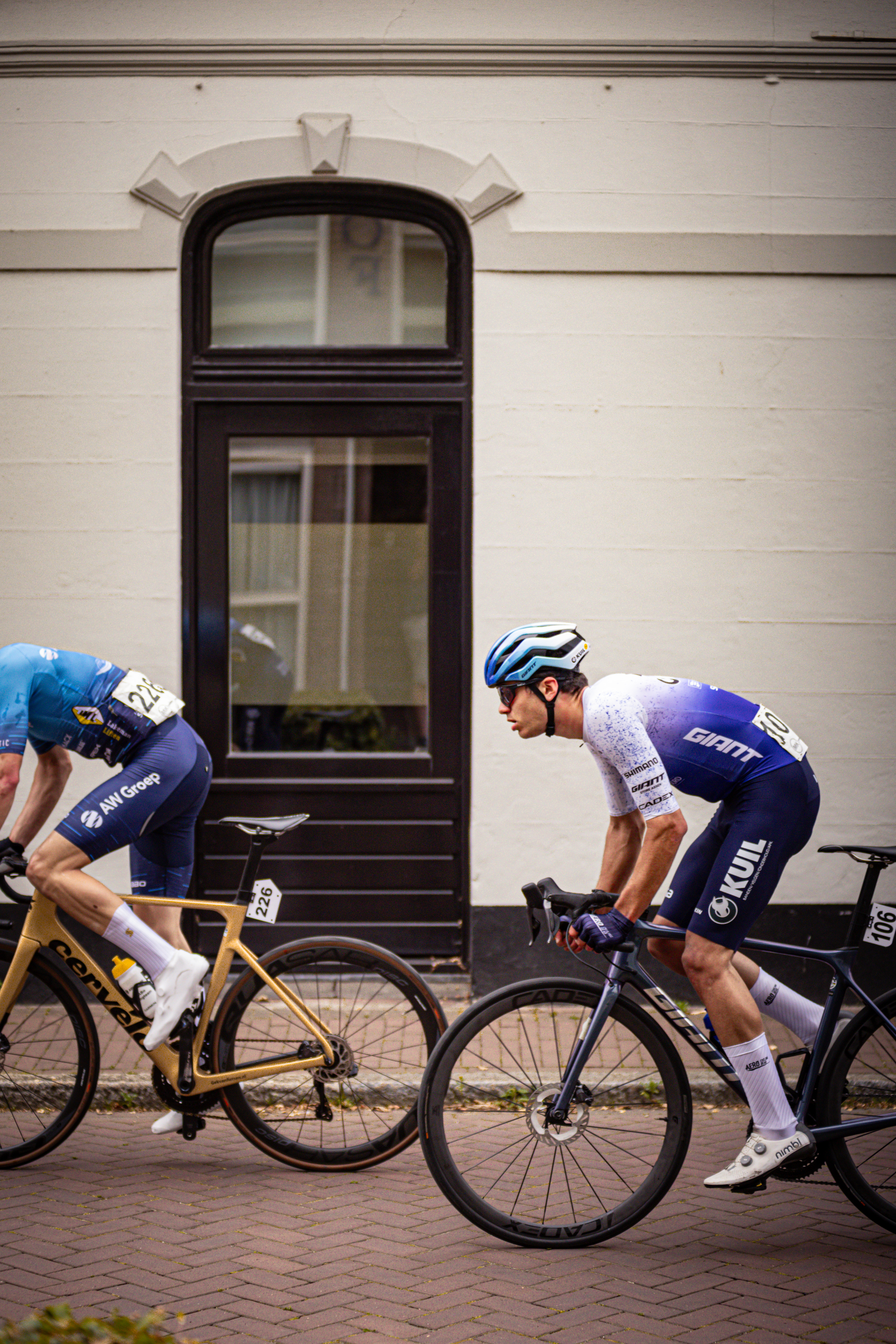 Two cyclists on a brick road in front of a building. One has the number 1 and the other 21 on his jersey.