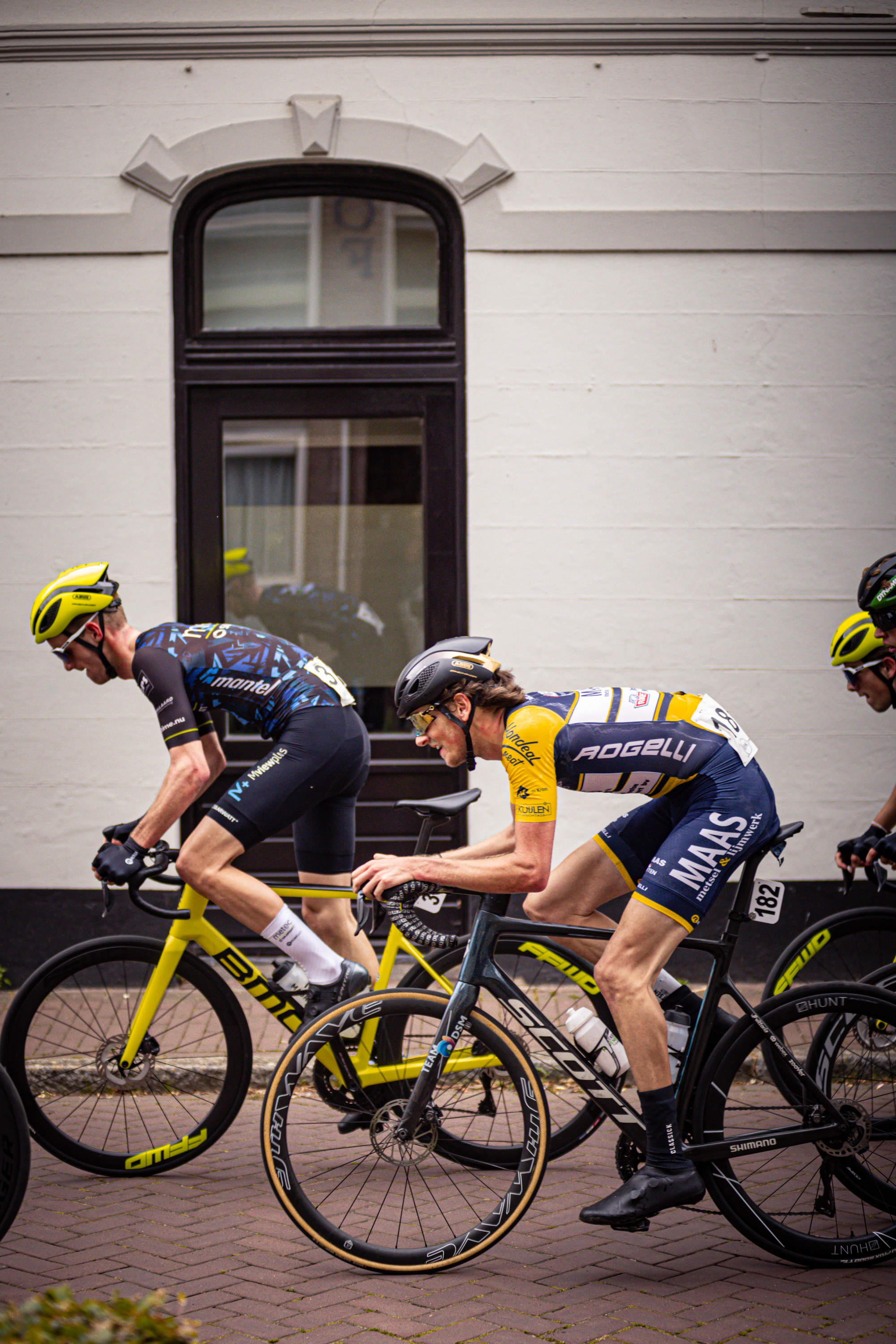 Three cyclists are seen wearing helmets and racing on a street near a building.