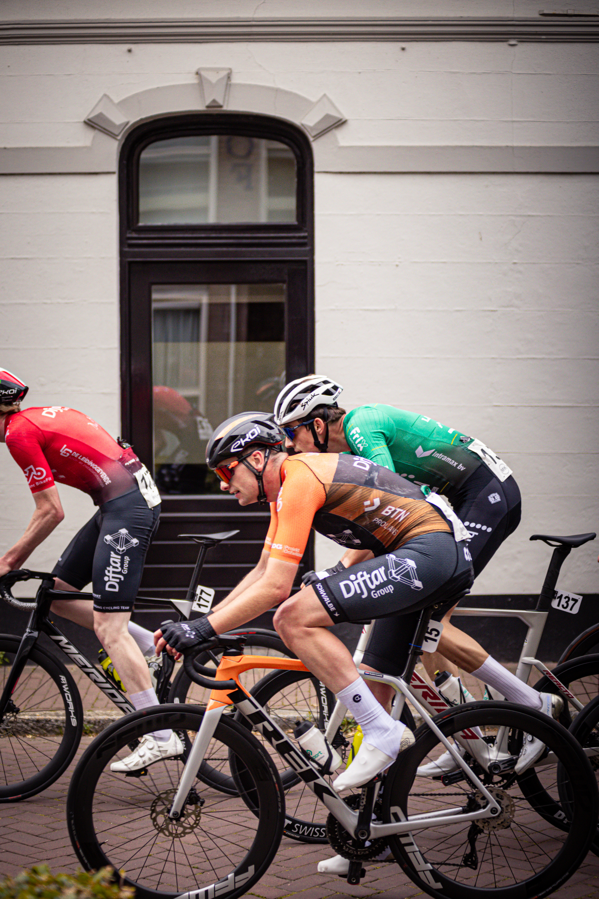 Three cyclists, one in a red jersey and two in black jerseys, race each other in front of a building with windows.