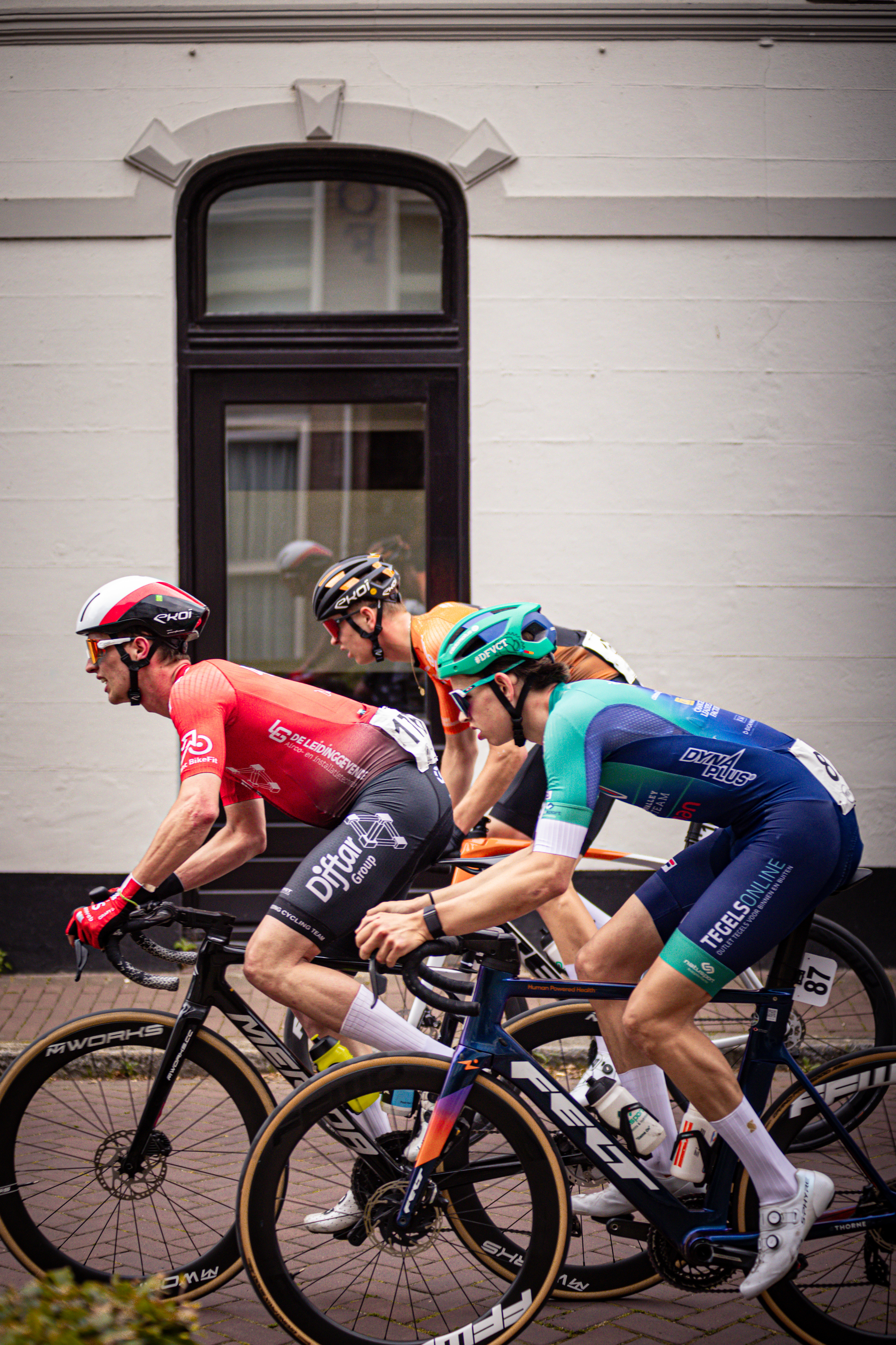 Three cyclists are riding in a line, with the leader wearing a blue and white jersey saying Wielrennen.