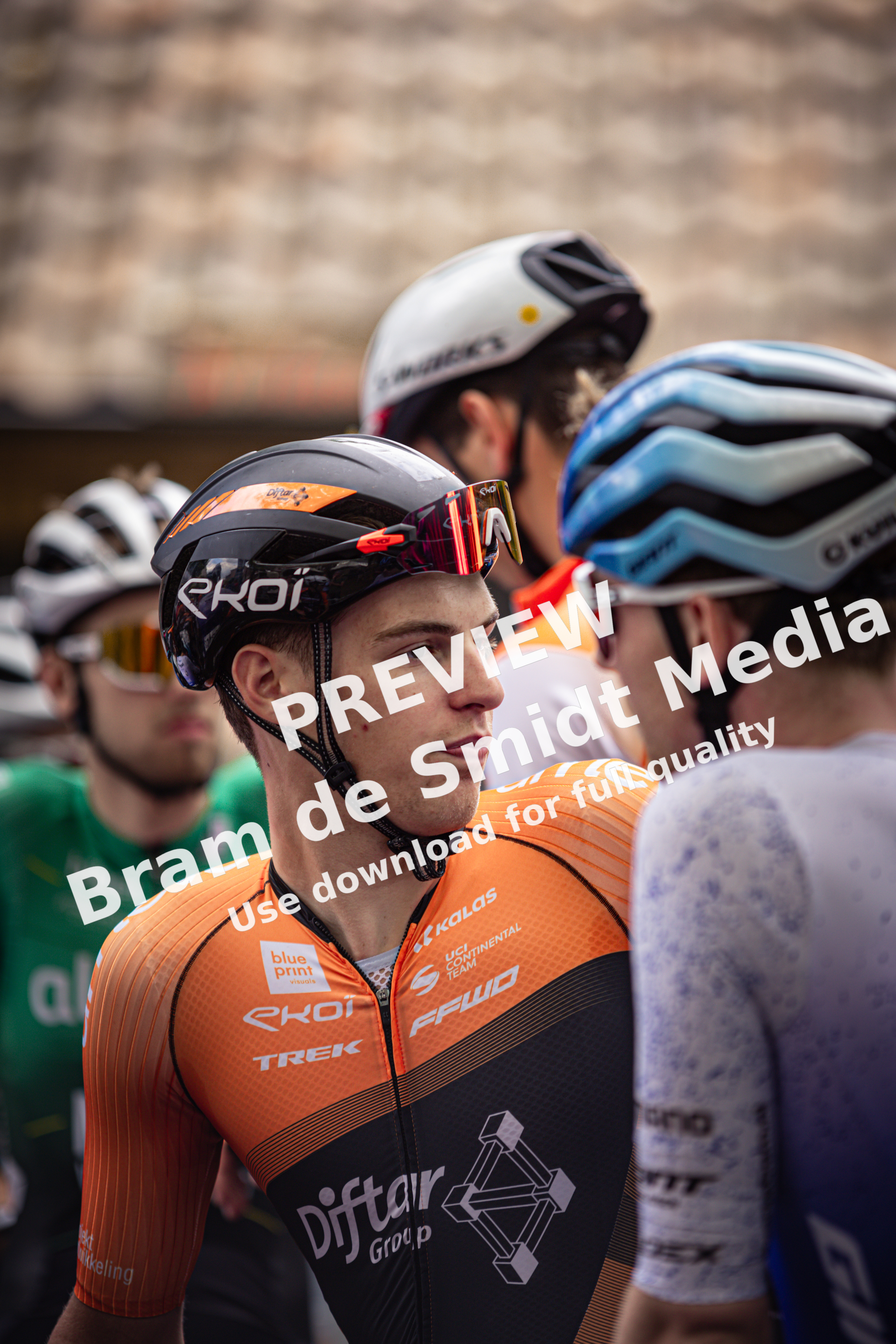 A group of cyclists in helmets, including a man wearing an orange jersey saying "Bouwmaat", standing on a street.