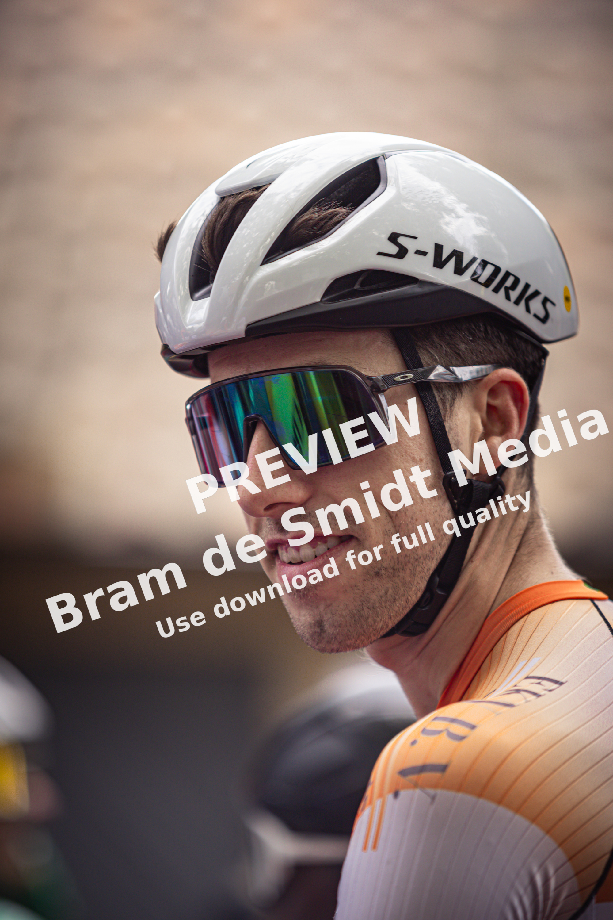 A cyclist wearing an orange and white jersey with the name Wielrennen on it, smiling at a photographer.