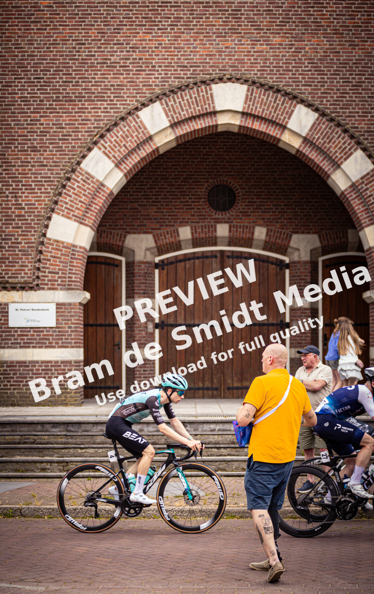 Two men on bicycles in front of a brick building that says "Midden Brabant Poort Omloop".