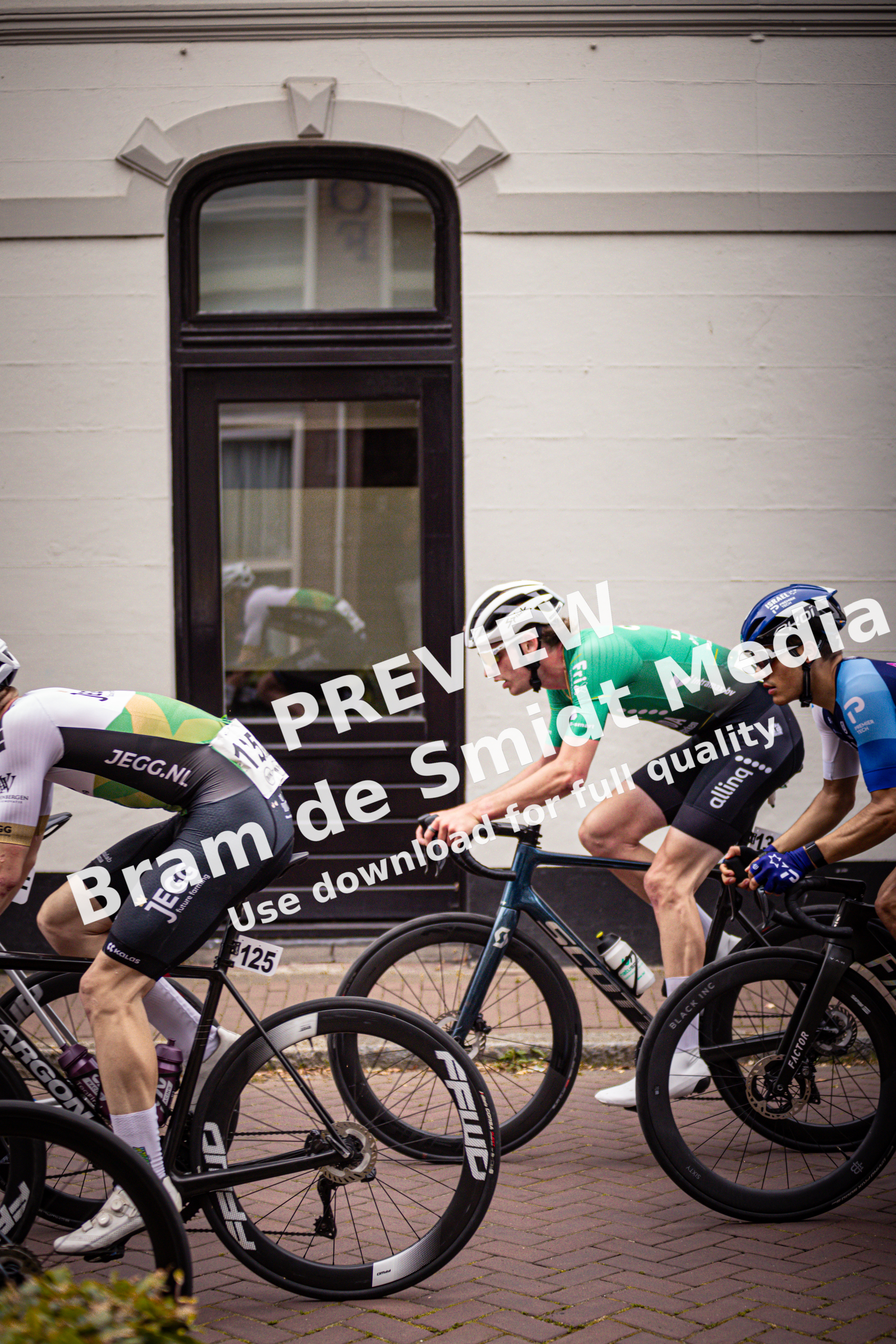 Four cyclists are riding past a building. The cyclist in the green jersey has his hand on his handlebars.