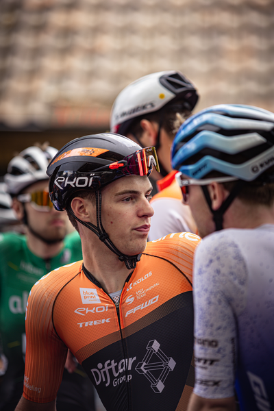 A group of cyclists in helmets, including a man wearing an orange jersey saying "Bouwmaat", standing on a street.