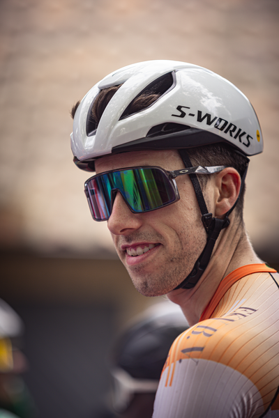 A cyclist wearing an orange and white jersey with the name Wielrennen on it, smiling at a photographer.