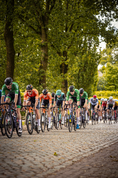 A group of cyclists riding down a cobblestone road, the leader is wearing an orange shirt.