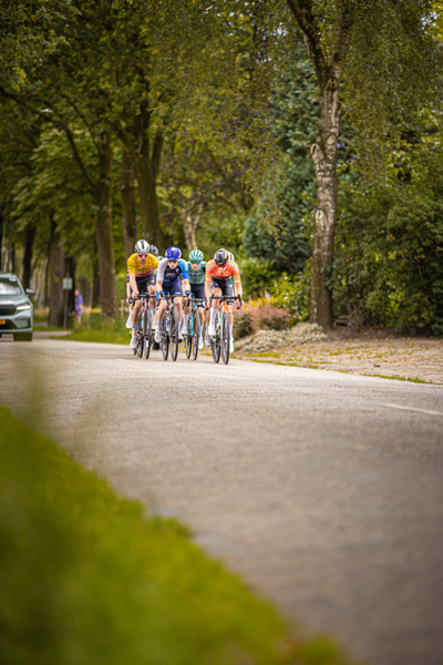 Four cyclists ride down a road in Midden Brabant, participating in the Poort Omloop race.