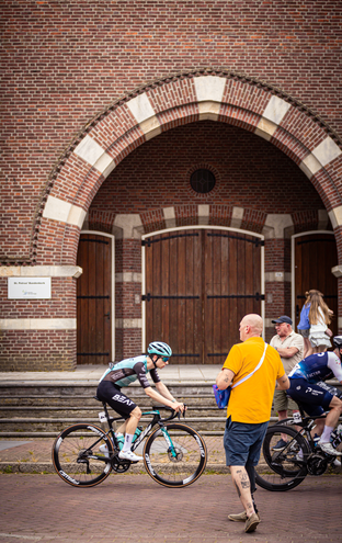 Two men on bicycles in front of a brick building that says "Midden Brabant Poort Omloop".