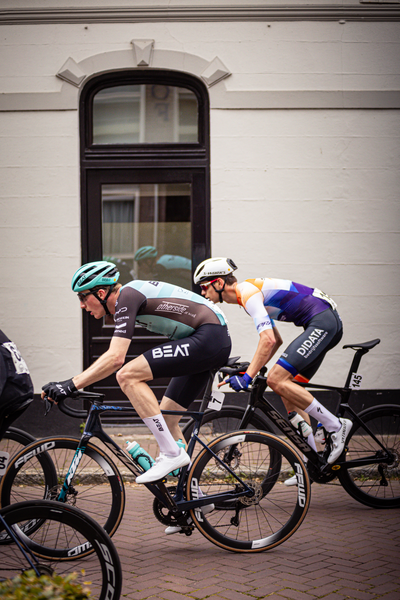 Two cyclists are in a race and they have bibs on that say "Beat". They are riding their bikes down a city street.