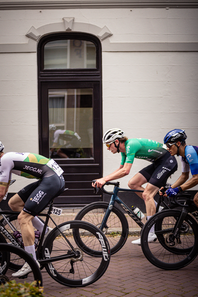 Four cyclists are riding past a building. The cyclist in the green jersey has his hand on his handlebars.