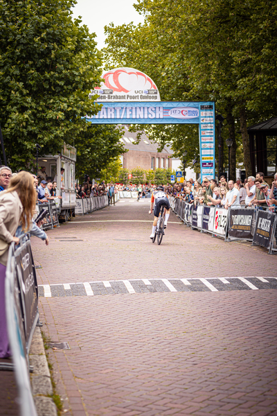 A cyclist in a blue and white uniform is riding past a crowd of people at the Midden Brabant Poort Omloop.