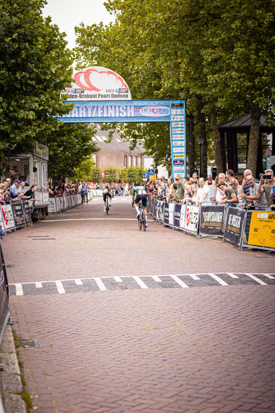 People watch a cyclist on a street lined with trees and buildings. The cyclist is wearing the number 2 on his jersey.